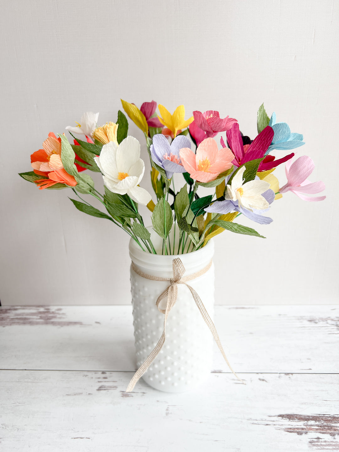 A colorful bouquet of paper wildflowers in shades of pink, yellow, orange, blue, and white, arranged in a textured white vase. The vase is decorated with a tied ribbon, adding a rustic charm. The bouquet sits on a light wooden surface against a plain background.