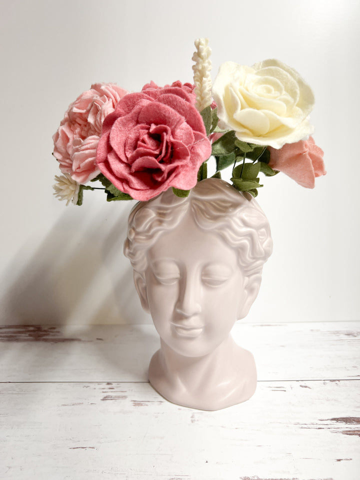 Alt text: A classical bust-shaped vase featuring a serene face, holding a felt flower bouquet with pink, cream, and red roses. The soft pastel colors of the flowers add a gentle contrast to the minimalist white surface and background, creating an elegant display.