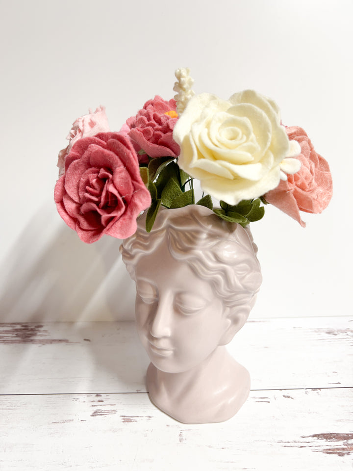 Alt text: A classical bust-shaped vase featuring a serene face, holding a felt flower bouquet with pink, cream, and red roses. The soft pastel colors of the flowers add a gentle contrast to the minimalist white surface and background, creating an elegant display.