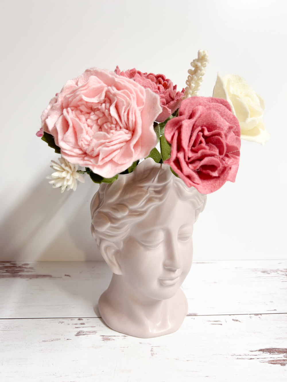 Alt text: A classical bust-shaped vase featuring a serene face, holding a felt flower bouquet with pink, cream, and red roses. The soft pastel colors of the flowers add a gentle contrast to the minimalist white surface and background, creating an elegant display.