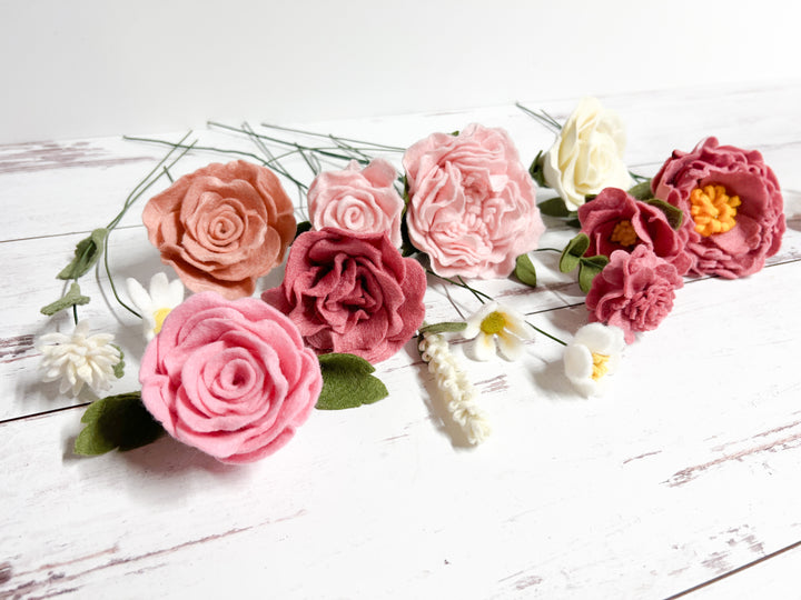 A collection of felt flowers, including pink, blush, cream, and burgundy roses, arranged on a white surface. The soft, textured petals and delicate colors create a charming, handcrafted aesthetic. Each flower has a long green stem, adding a natural touch to the display.